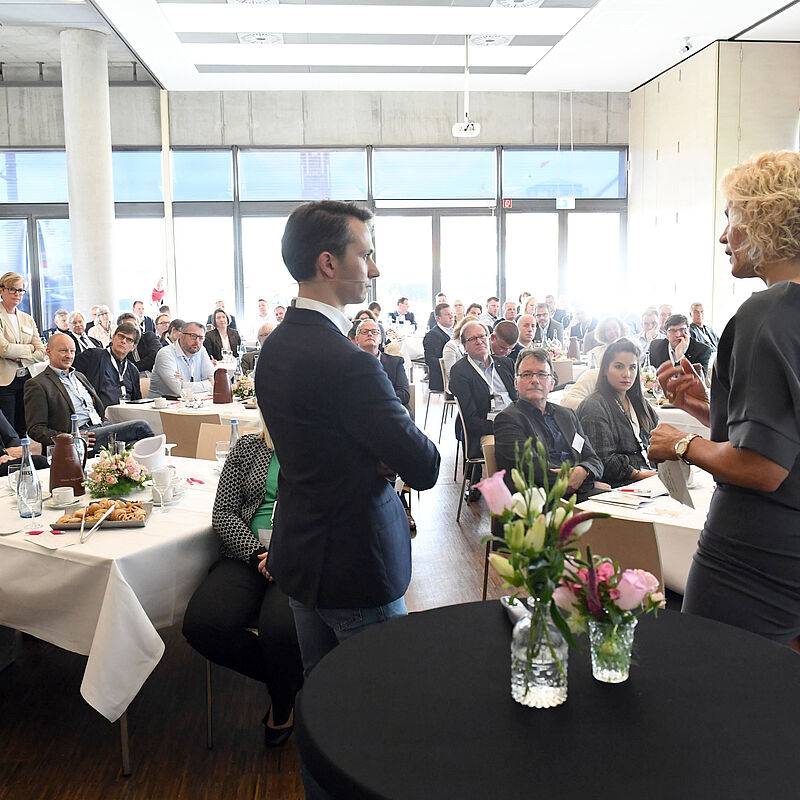 Redner und Moderatorin stehen gemeinsam auf der Bühne, davor eine gespannte Zuhörerschaft an Tischen