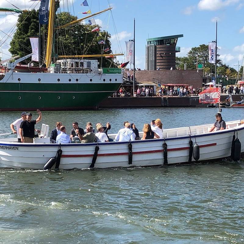 Eine Menschengruppe auf einer Bootstour in einem kleinen Motorboot mit Blick auf größere Segelschiffe im Hintergrund