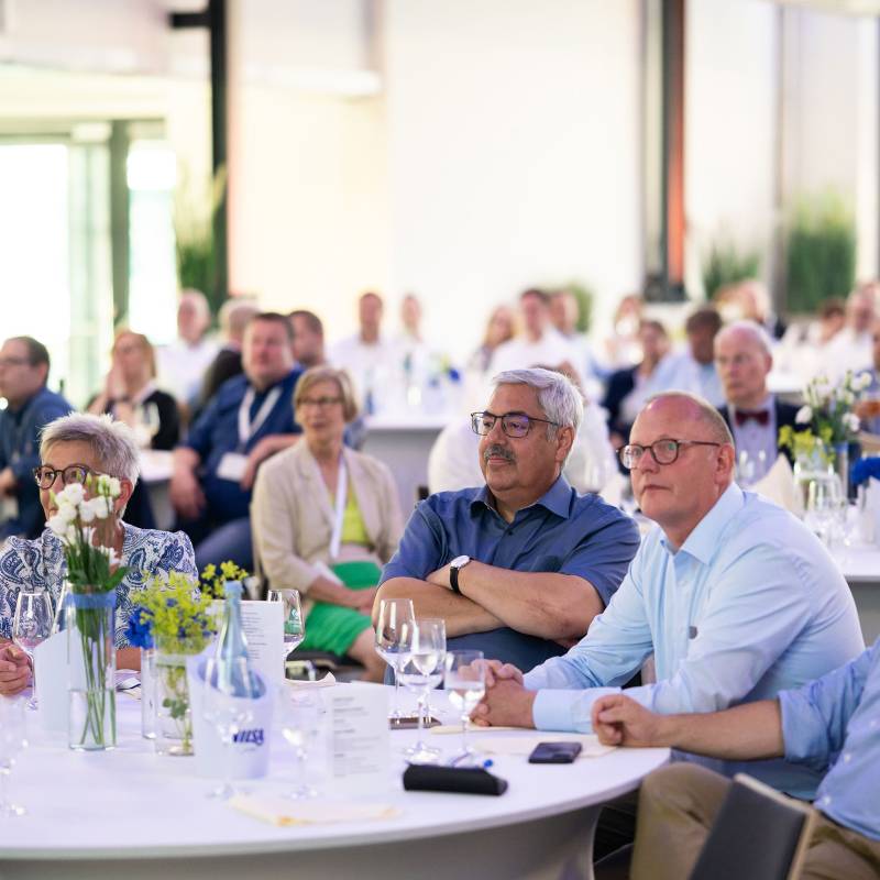 Bürgermeister Melf Grantz bei der Eröffnungsveranstaltung. Abgebildet ist ein Raum voller Menschen mit zwei Männern mittleren Alters im Fokus, beide tragen eine Brille der rechte hat eine Glatze.